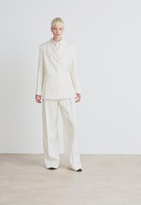 Completo bianco a righe sottili con giacca doppiopetto su misura e pantaloni a gamba larga. Tessuto morbido con linee nette, completato da scarpe nere.