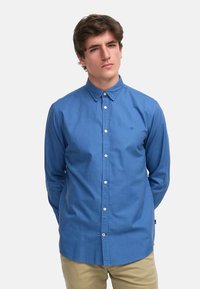 Camisa azul de botón con cuello puntiagudo, mangas largas y un pequeño logotipo bordado. Fabricada con una tela texturizada.
