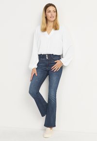 Valkoinen, pitkähihainen paita V-pääntieellä yhdistettynä korkeavyötäröisiin, leveneviin farkkuihin tummassa pesussa. Vaaleat liukkaat kengät.