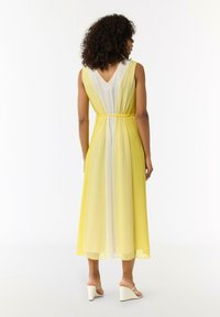 Gelb-weißer gradient Maxi-Kleid mit V-Ausschnitt, tailliertem Bund und transparentem Stoff, kombiniert mit weißen Wedge-Heels.