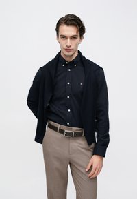 Chemise bleu marine boutonnée avec un pull foncé posé sur les épaules, associée à un pantalon beige clair et une ceinture marron. Design simple et épuré.