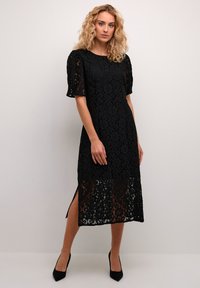 Black lace dress with floral pattern, short puff sleeves, side slits, and a fitted silhouette, paired with black high heels.