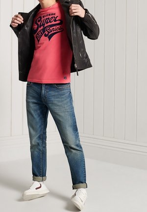 Sort læderjakke med lynlås, pink grafisk t-shirt med marineblå tekst, blå jeans og hvide sneakers med røde detaljer, stående på en lys gulv.