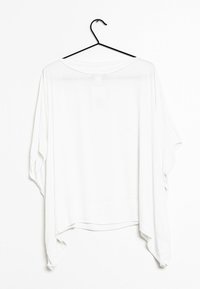 Top bianco, leggero, dal taglio ampio e maniche a batwing. Presenta un orlo curvo e una texture liscia, appeso a un gancio nero.