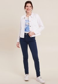 Witte denimjas met kraag, zilveren knopen en twee voorzakken, gecombineerd met een blauw t-shirt met wolkenpatroon en donkere skinny jeans.