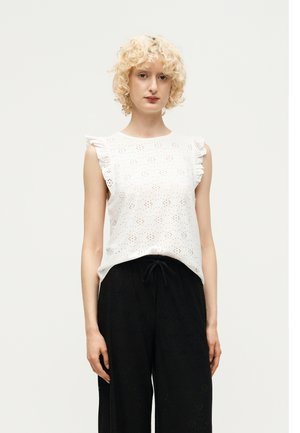 Ung person med krøllet blondt hår iført hvid ærmeløs øjetsbluse med flæsekant og sorte vide bukser med snøre.