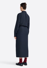 Lungo abito blu navy con righe verticali, design con collo, maniche lunghe e vita stretta. Abbinato a calze rosse e scarpe nere.