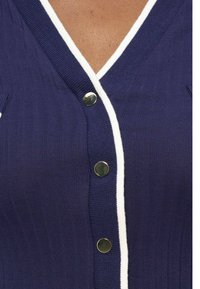 Κοντινό πλάνο μιας μπλε ναυτικού χρώματος πλεκτής ζακέτας με λευκή επένδυση και τρία στρογγυλά χρυσά κουμπιά, φορεμένη πάνω σε ανοιχτό καφέ τόνο δέρματος.