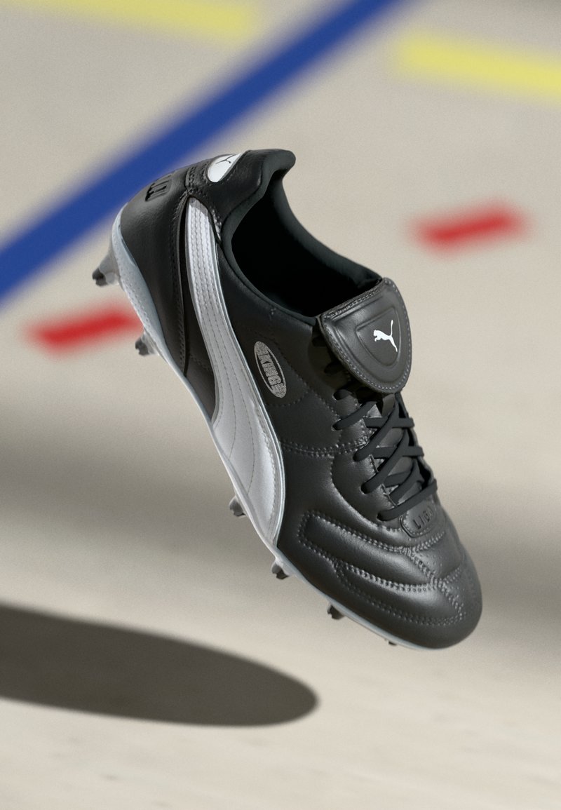Black and white Puma soccer cleat with laces and studs, floating above a light surface with colored lines in the background.