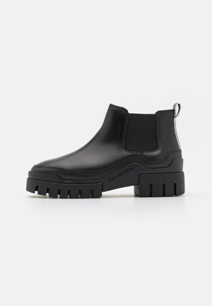 Botte Chelsea en cuir noir avec semelle à crampons épaisse, panneau latéral élastique, marquée "Calvin Klein Jeans", et languette de traction sur fond blanc.