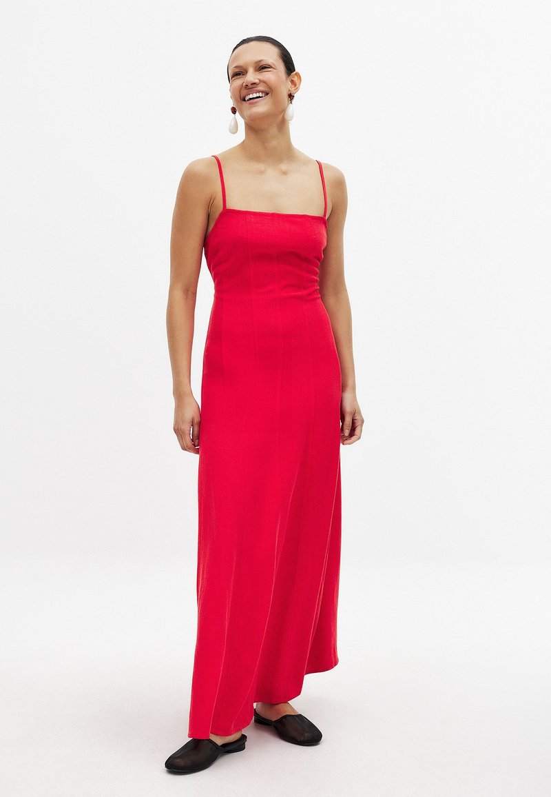 Röd maxiklänning med tunna axelremmar, en figurnära överdel och en flödande kjol. Bärs med svarta slip-on skor. Slät textur, minimalistisk design.