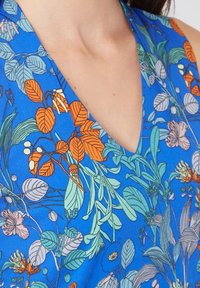 Primer plano de una prenda azul con patrones florales en naranja, verde y pálido, y un escote en V que muestra parte del cuello y el hombro de una persona.