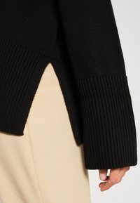 Svart stickad tröja med ribbade ärmslut och sidoflikar, som visar en texturerad tyg. Buren över ljusbeiga byxor.