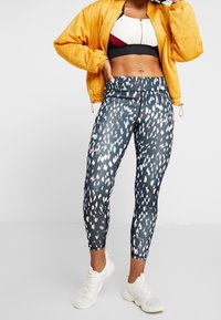 Leggings en un tejido oscuro y estampado que presenta acentos en blanco y azul claro. Combinados con un top deportivo corto en blanco y negro y una chaqueta bomber amarilla.