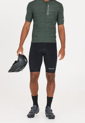 Man die een groen fietsshirt met korte mouwen en zwarte gevoerde korte broek draagt, houdt een zwarte fietshelm in één hand.