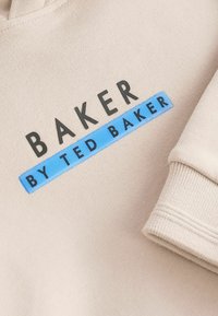 Sudadera beige con el texto negro en relieve "BAKER" y una etiqueta azul con "by TED BAKER". Tejido suave con puños acanalados.