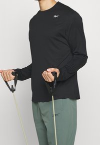 Svart långärmad träningströja med rundad hals och en vit logotyp, ihop med gröna byxor, händer som greppar motståndsband.