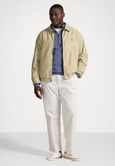 Beige lichtgewicht jack over een blauwe pullover, gecombineerd met witte broeken en witte sneakers. Het jack heeft een kraag en een rits.