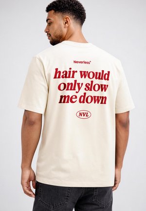 Man with short curly hair wearing a cream t-shirt with red text on the back reading "hair would only slow me down" and black pants.