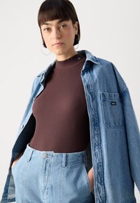 Brauner gerippter Rollkragenpullover unter einem hellblauen Denimhemd mit einer Brusttasche. Das Hemd hat Metallknöpfe und ein kleines Logodetail.