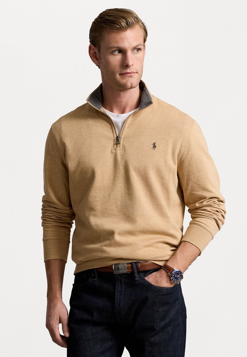 Beige kvartdragningströja med en grå krage, tillverkad av mjukt material, med en liten logotyp på bröstet, bärs tillsammans med mörka jeans.