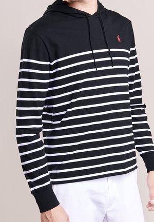 Homme portant un sweat à capuche noir avec des rayures horizontales blanches et un logo rouge sur la poitrine, associé à un pantalon blanc, sur un fond uni.