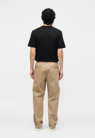 Άνδρας με σγουρά μαλλιά που φοράει μαύρο μπλουζάκι και μπεζ παντελόνι, στέκεται γυρισμένος με την πλάτη προς ένα απλό φωτεινό φόντο.