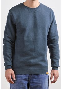 Sudadera azul oscura de tela suave, con puños y dobladillo acanalados, cuello redondo y un pequeño parche de logo en la manga izquierda.