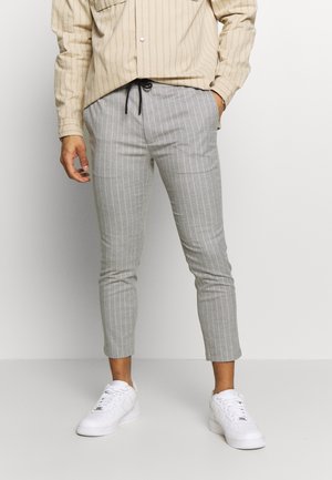 Mann trägt graue, verkürzte Nadelstreifenhose mit schwarzem Kordelzug, beiges gestreiftes Hemd und weiße Turnschuhe, steht vor einer einfachen hellen Wand.