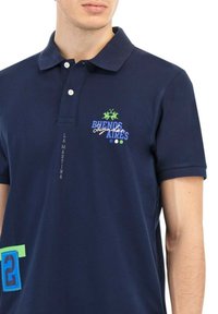 Uomo che indossa una polo blu notte con il testo "Buenos Aires", un logo di un giocatore di polo verde e una toppa blu con il numero 2 sul lato inferiore.