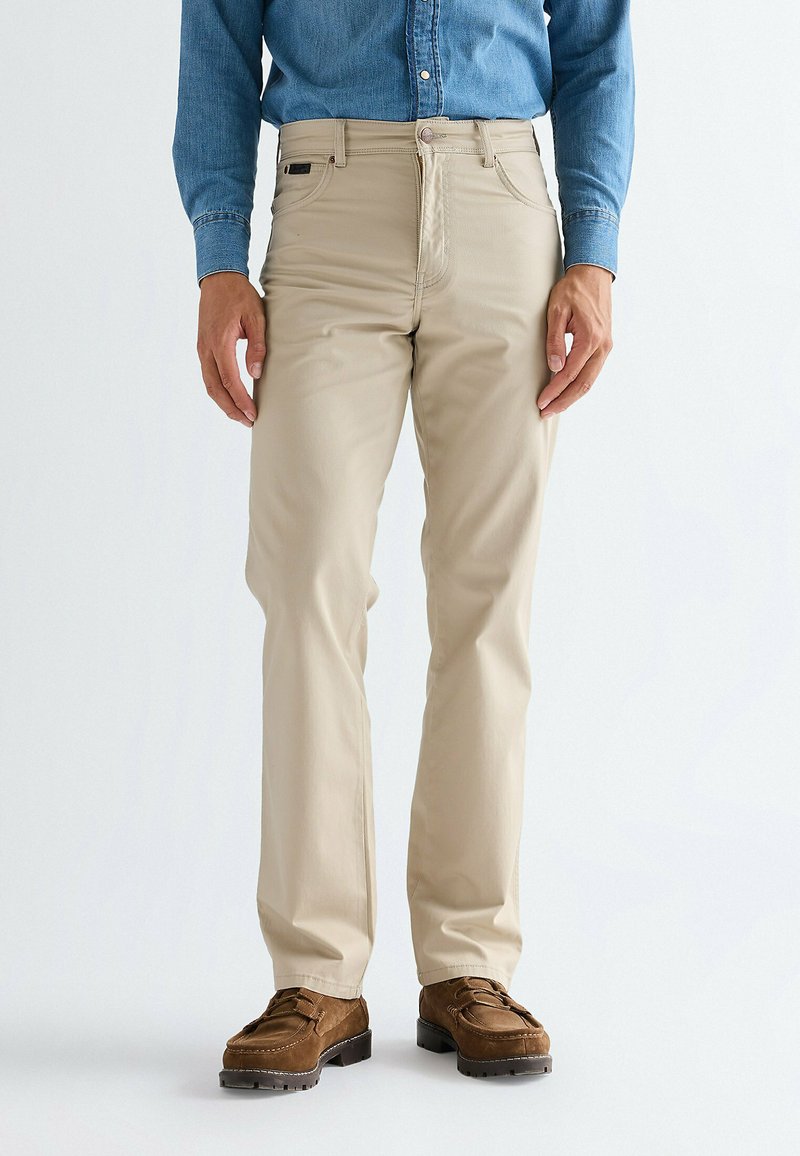 Hombre con pantalones beige, zapatos de ante marrones y una camisa de mezclilla azul, de pie frente a un fondo blanco liso.