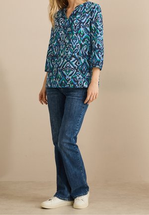 Kobieta ubrana w bluzkę w niebiesko-zielono-czarne wzory z rękawami do łokcia, ciemnoniebieskie dżinsy i białe trampki stoi przy beżowej ścianie.