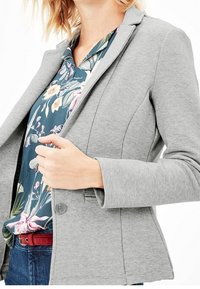 Blazer grigio con spalle strutturate, chiusura a bottone e tessuto testurizzato, indossato sopra una blusa a fantasia floreale. Stile casual ma curato.