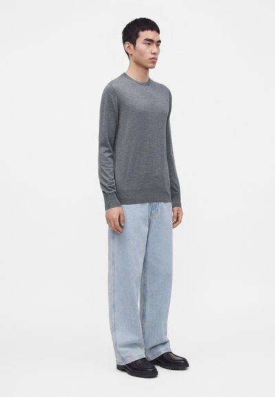 Jeune homme debout portant un pull gris à manches longues, un jean bleu clair à jambes larges et des chaussures noires sur un fond blanc uni.