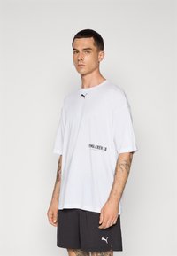 Hvit t-skjorte med rund hals og korte ermer, med en svart Puma-logo og teksten "PMA CREW 48" på forsiden. Mykt materiale.
