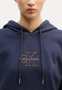 Moleton azul-marinho com cordões, apresentando um logótipo bordado em laranja "Calvin Klein" e "CK" numa fonte elegante no peito.