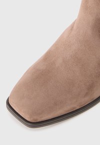 Botine din suede de culoare bej deschis, cu vârf pătrat și textură fină. Prezintă o talpă subțire neagră pentru contrast.