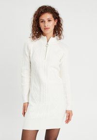 Robe tricotée blanche avec un col zippé mi-hauteur, présentant des motifs en relief de type câble et des poignets côtelés. Tissu lisse avec une silhouette ajustée.