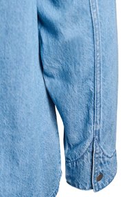 Svetlo modrý denimový rukáv košele s viditeľným prešitím švov a cvokovým gombíkom na manžete na bielom pozadí.