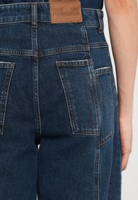 Tumši ziloņkaulu pārvalki ar teksturētu apdari, ar vairākiem kabatām un kontrastējošiem oranžiem šuvumiem. Brūna etiķete ar zīmola nosaukumu aizmugurē.