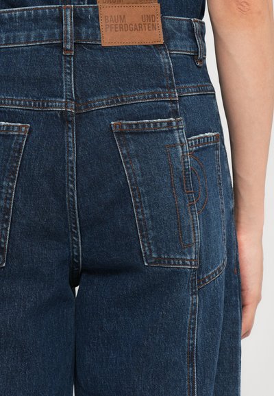Donkerdenim dungarees met een gestructureerde afwerking, voorzien van meerdere zakken en contrasterende oranje stiksels. Bruin label met merknaam op de achterkant.