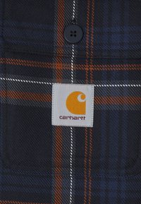 Närbild på ett marinblått och orange rutigt tyg med en svart knapp och en vit Carhartt-logopatch sydd på tyget.