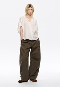 Helles beige, kurzärmliges Hemd mit Knöpfen, kombiniert mit locker sitzenden, olivgrünen Hosen aus strukturiertem Stoff und weitem Schnitt.