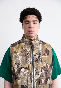 Camo-mønstret vest i brun, grøn og beige med en frontlynlås, to forlommer og Adidas-logo i rød. Grøn skjorte med korte ærmer under.