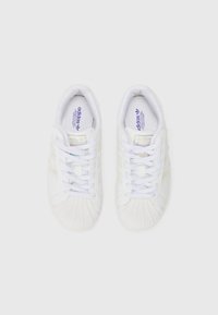 Baskets de sport blanches avec une coque de protection texturée, dessus en tissu, rayures embossées et un branding contrasté sur la languette et la semelle intérieure.