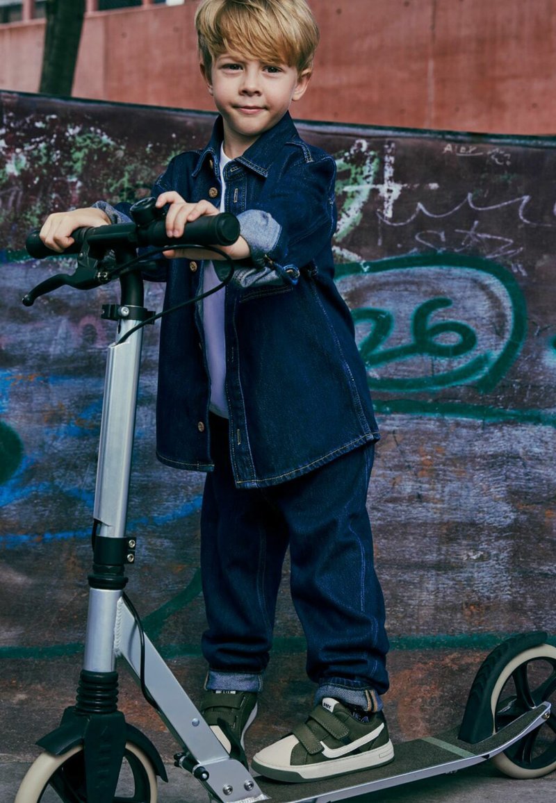 Jeune garçon portant une veste en jean et un pantalon, debout avec un pied sur une trottinette devant un mur couvert de graffitis.