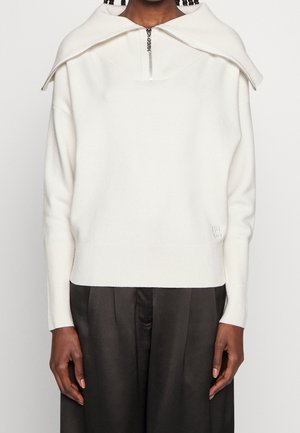 Persoon draagt een witte gebreide trui met een halve ritssluiting en een omgeslagen kraagdetail, gecombineerd met losse zwarte broek.