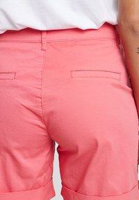 Różowe bawełniane szorty o gładkiej fakturze, z dwiema tylnymi kieszeniami i mankietami. Brak widocznych elementów metalowych lub wzorów.