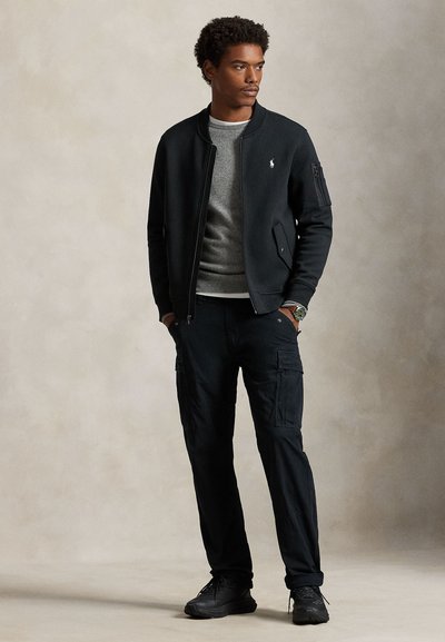 Mees kannab musta bomberjopet halli kampsuni peal, mustad cargo-püksid ja mustad tossud, seisab käed taskutes.