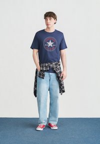 Νεαρός άνδρας στέκεται με τα χέρια στις τσέπες, φορώντας μπλε κοντομάνικο μπλουζάκι Converse All Star, ανοιχτό μπλε τζιν, κόκκινα αθλητικά παπούτσια και καρό πουκάμισο δεμένο γύρω από τη μέση.